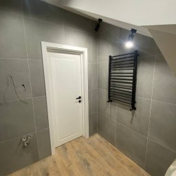 DK Renovations Homes - Wykończona łazienka z szarymi płytkami, drewnopodobną podłogą, białymi drzwiami z czarną klamką i czarnym grzejnikiem drabinkowym oraz dwoma reflektorami punktowymi.