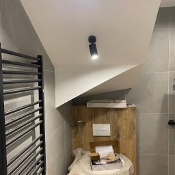 DK Renovations Homes - Nowoczesna łazienka w szarościach z drewnianą zabudową podtynkową toalety, czarnym grzejnikiem drabinkowym i punktowym oświetleniem sufitowym; widoczne opakowania i elementy montażowe toalety przed...