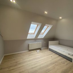 DK Renovations Homes - Poddasze po remoncie: jasne panele podłogowe, szara ściana do połowy, biała góra, dwa okna dachowe, biały grzejnik, białe łóżko z szarymi szufladami.