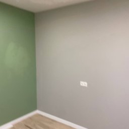 DK Renovations Homes - Pokój po świeżym malowaniu: jedna ściana w odcieniu zieleni, druga szara, widoczne gniazdko elektryczne, podłoga wyłożona panelami.