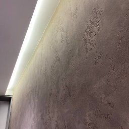 DK Renovations Homes - Teksturowana ściana w odcieniach brązu z subtelnym oświetleniem LED wzdłuż sufitu, ukazująca detale wykończenia i nieregularną fakturę powierzchni.