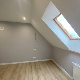 DK Renovations Homes - Poddasze po remoncie: świeżo pomalowane szare ściany, drewnopodobna podłoga, okno dachowe z drewnianą ramą i biały grzejnik.