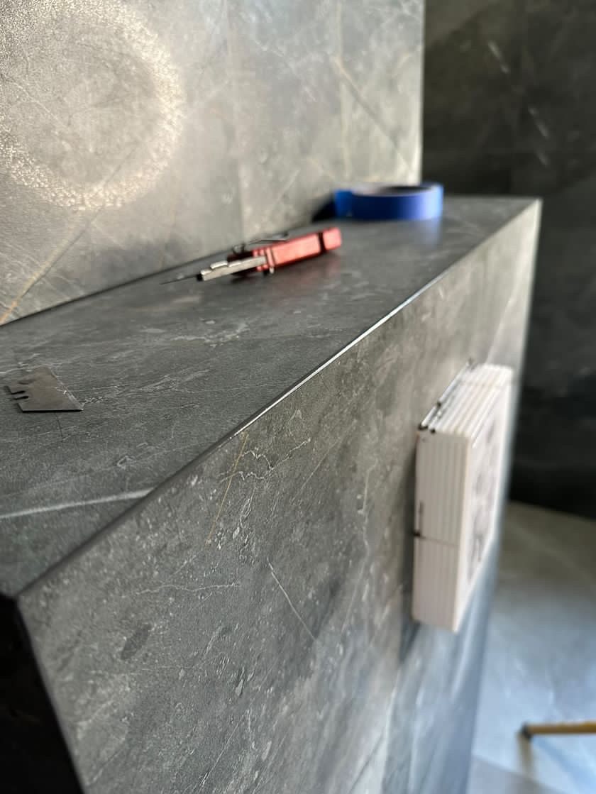 Szara, marmurkowa półka w trakcie montażu z narzędziami: nożyk z czerwoną rączką, niebieska taśma malarska i biały dozownik na ręczniki papierowe.