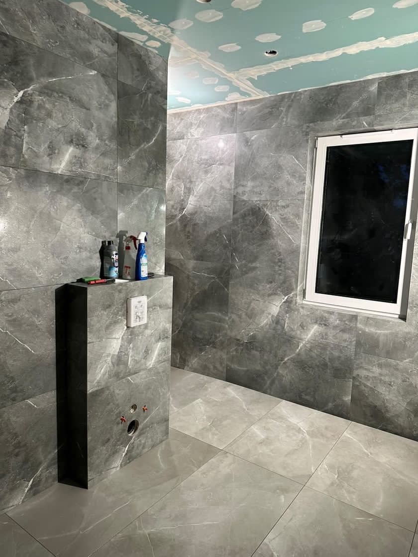 Łazienka w trakcie wykańczania z szarymi płytkami imitującymi marmur na ścianach i podłodze, widoczne przyłącza hydrauliczne do toalety podtynkowej, sufit z płyt kartonowo-gipsowych przed...