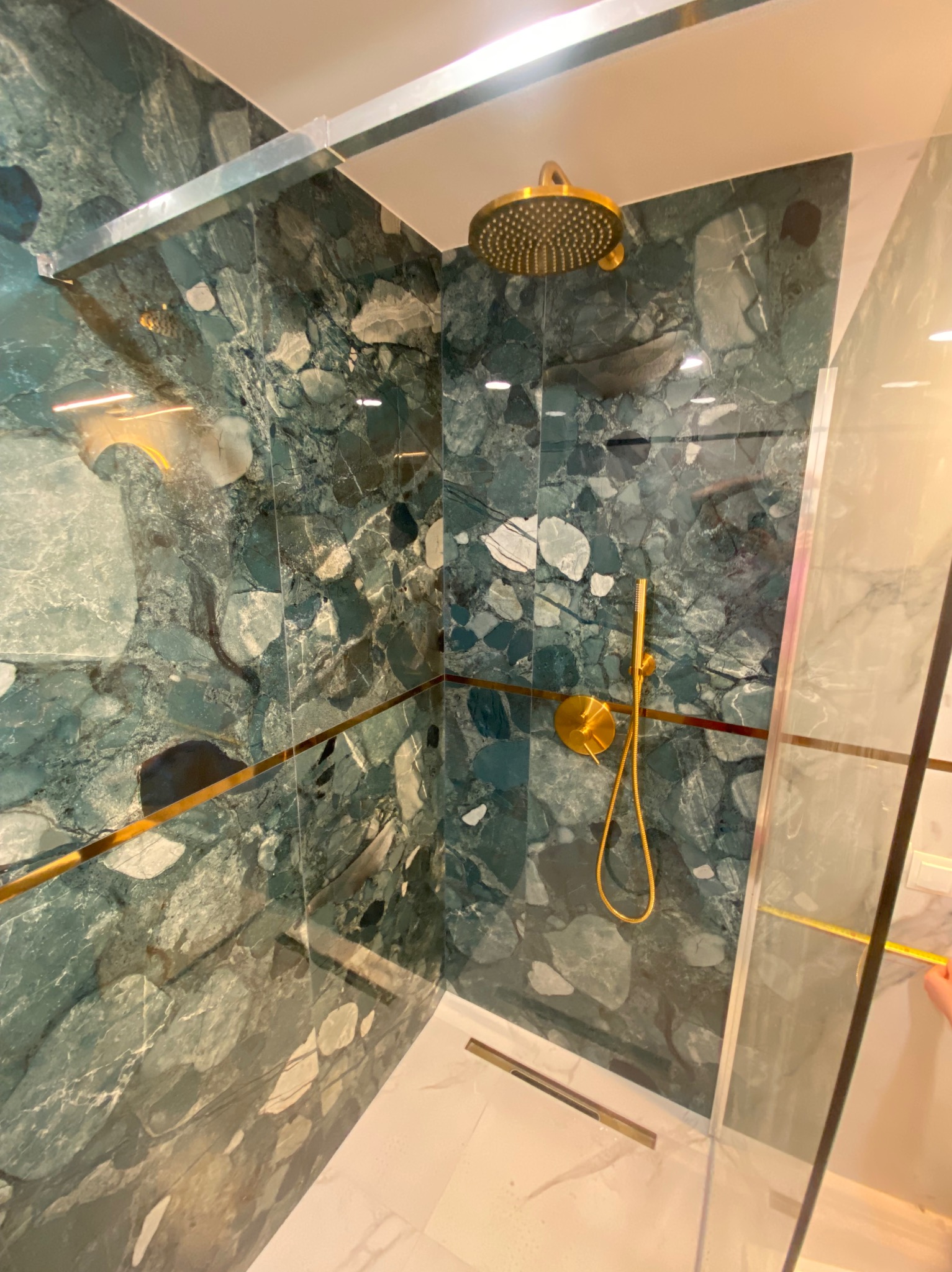 Kabina prysznicowa z zielonymi płytami imitującymi kamień z złotymi akcentami, deszczownica i bateria w kolorze złotym, biała podłoga.