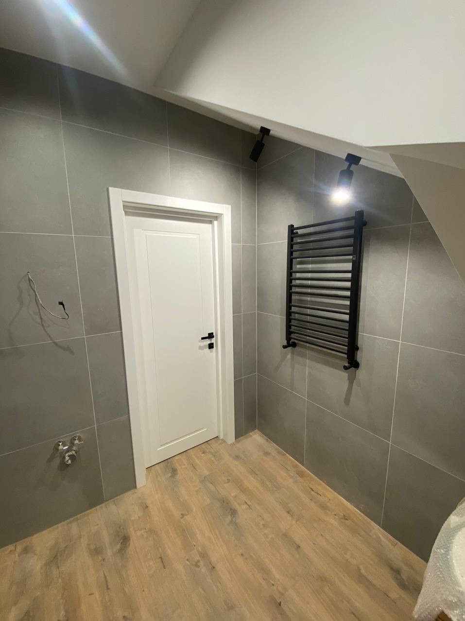Wykończona łazienka z szarymi płytkami, drewnopodobną podłogą, białymi drzwiami z czarną klamką i czarnym grzejnikiem drabinkowym oraz dwoma reflektorami punktowymi.