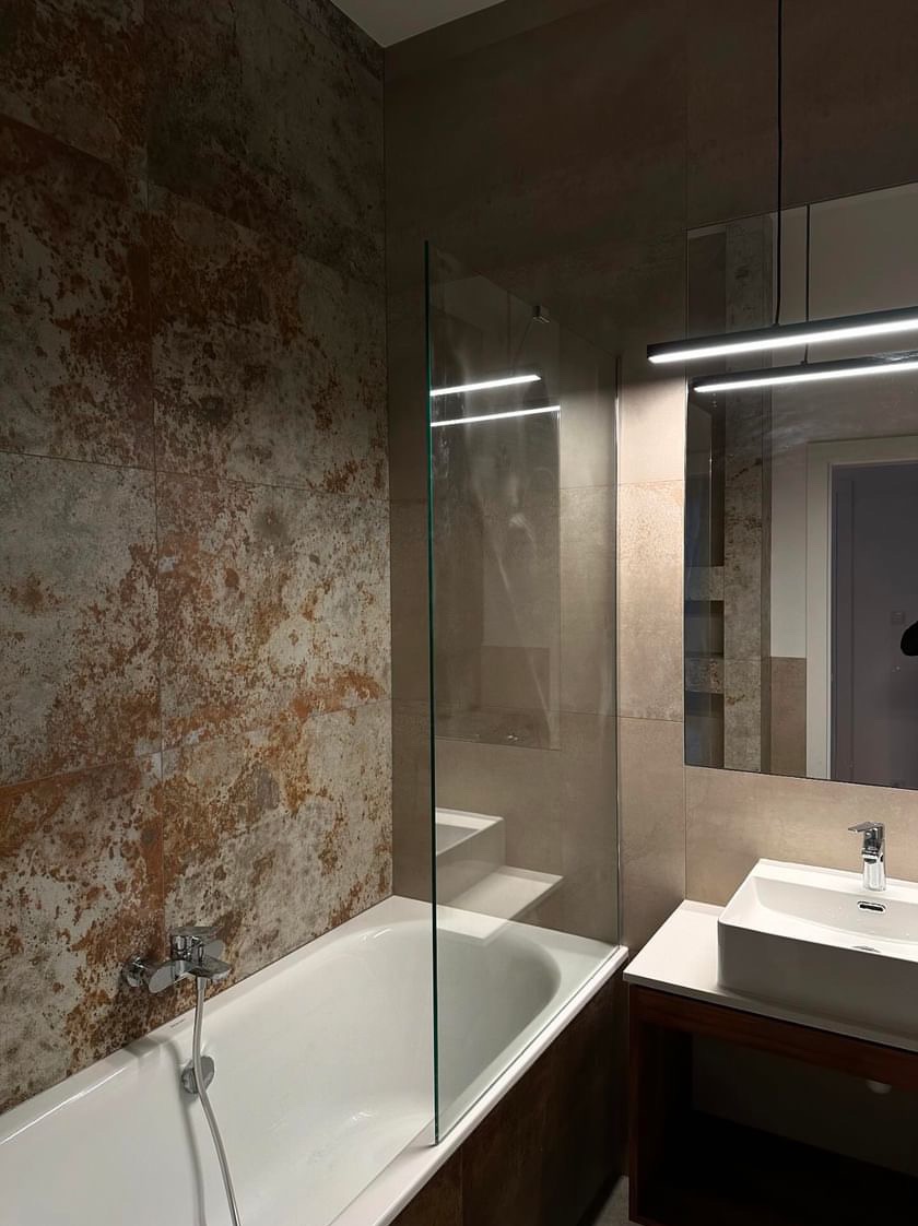 Nowoczesna łazienka z wanną, szklanym panelem prysznicowym i umywalką nablatową, płytki imitujące rdzę na ścianie, oświetlenie LED.