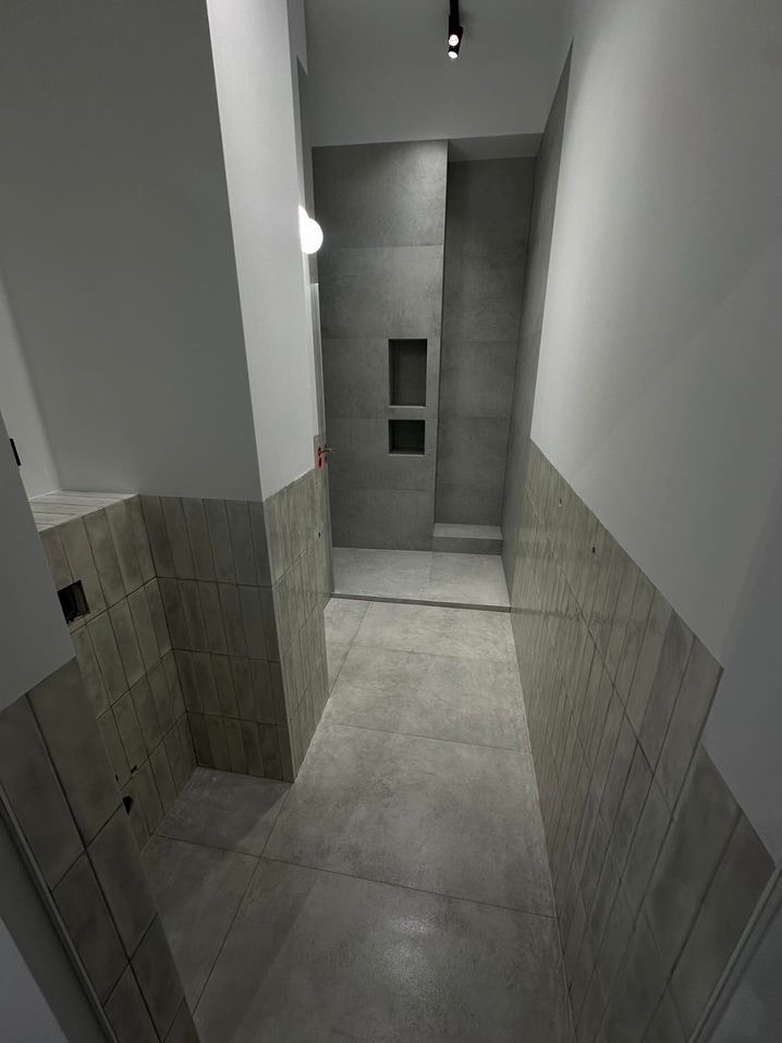 Szara łazienka z płytkami na ścianach i podłodze, widoczne wnęki w ścianie prysznicowej, minimalistyczny design i oświetlenie punktowe.