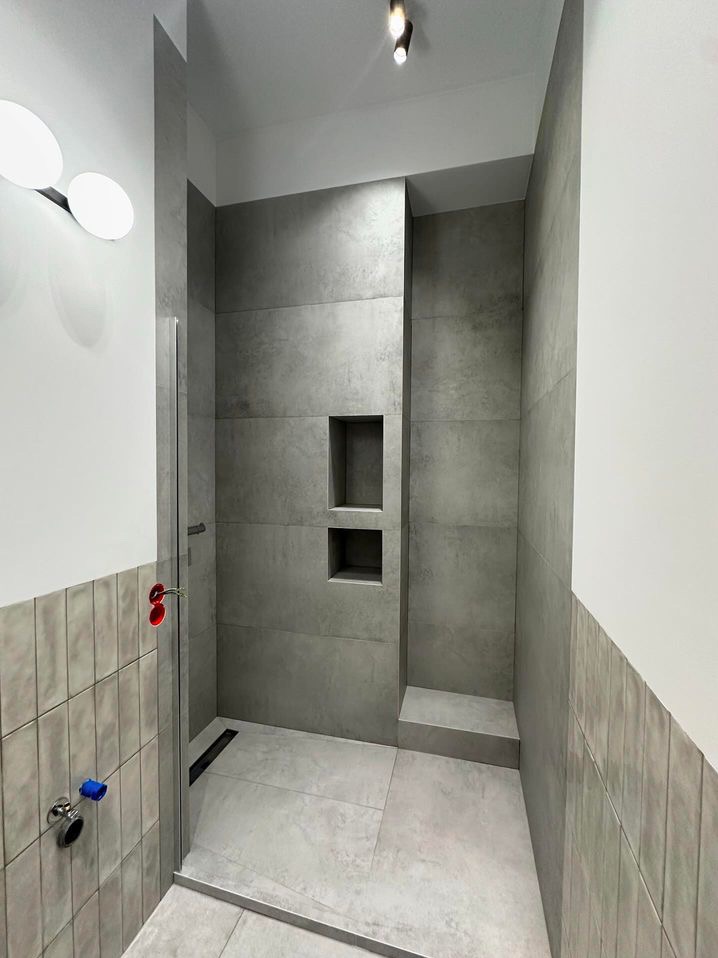 Nowoczesna kabina prysznicowa z szarymi płytkami, wnękami na kosmetyki i niskim progiem, widoczne przyłącza wody z czerwonym i niebieskim zaworem.