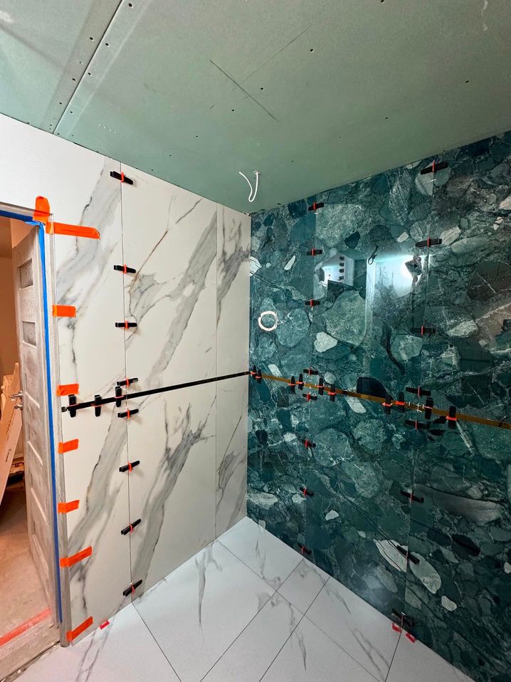 Układanie płytek marmurowych w łazience, widoczne płytki w odcieniach bieli i zieleni, system poziomowania płytek z pomarańczowymi klinami, surowy sufit z płyt kartonowo-gipsowych.