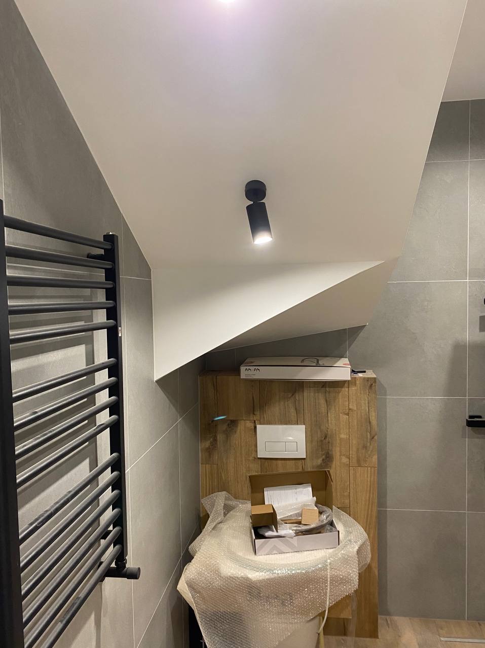 Nowoczesna łazienka w szarościach z drewnianą zabudową podtynkową toalety, czarnym grzejnikiem drabinkowym i punktowym oświetleniem sufitowym; widoczne opakowania i elementy montażowe toalety przed...