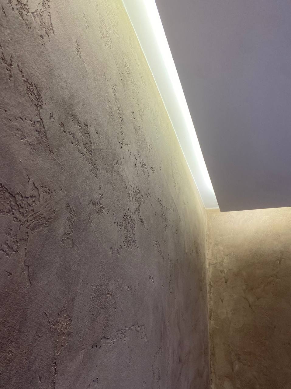 Ściana z dekoracyjną, strukturalną powierzchnią w odcieniach brązu i beżu, oświetlona listwą LED przy suficie.