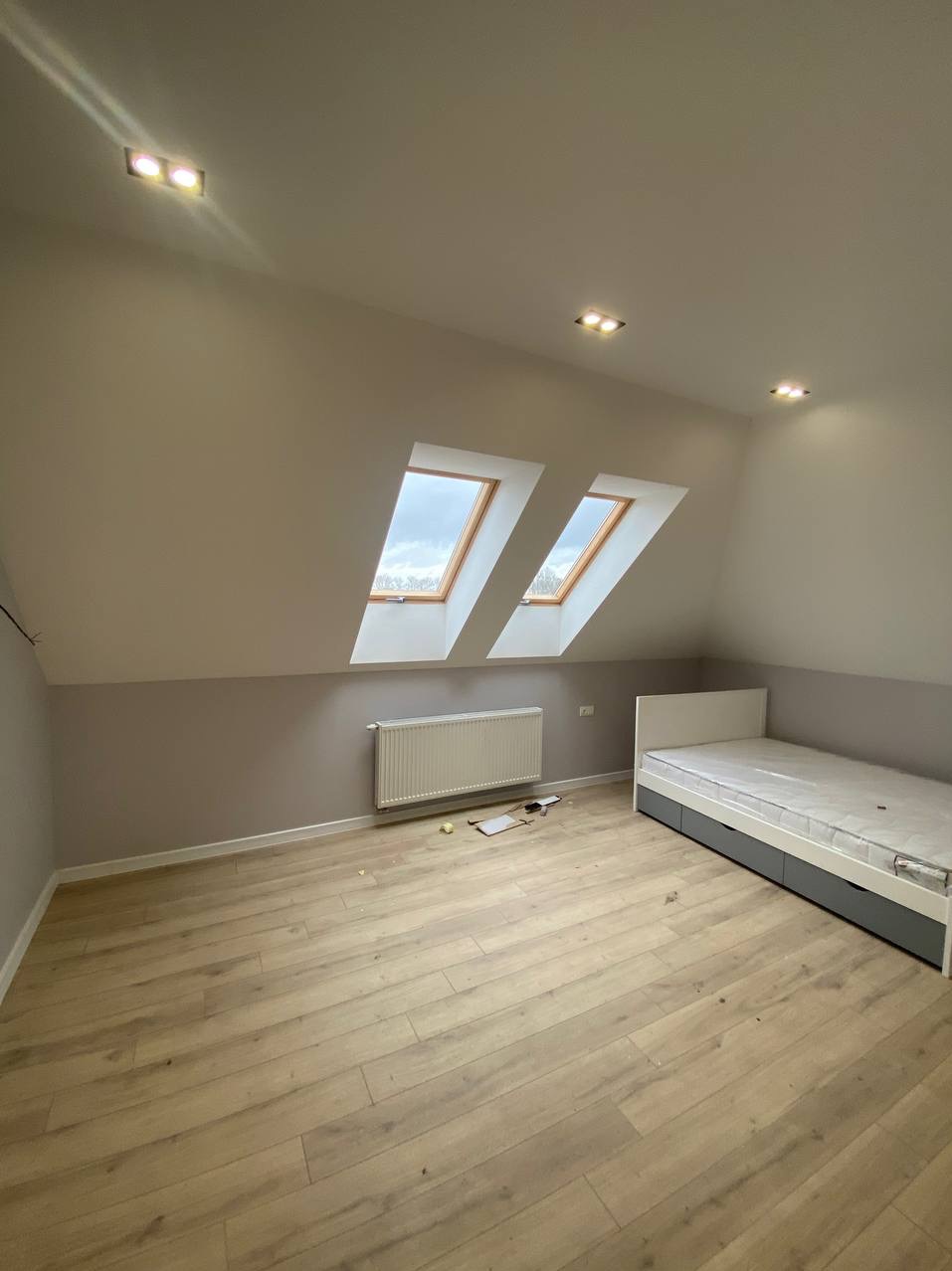 Poddasze po remoncie: jasne panele podłogowe, szara ściana do połowy, biała góra, dwa okna dachowe, biały grzejnik, białe łóżko z szarymi szufladami.