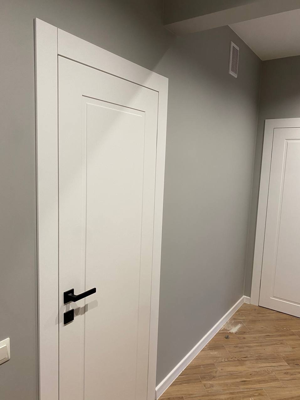 Białe, nowocześnie wykończone drzwi z czarną klamką na tle szarej ściany z listwą przypodłogową i drewnopodobną podłogą.