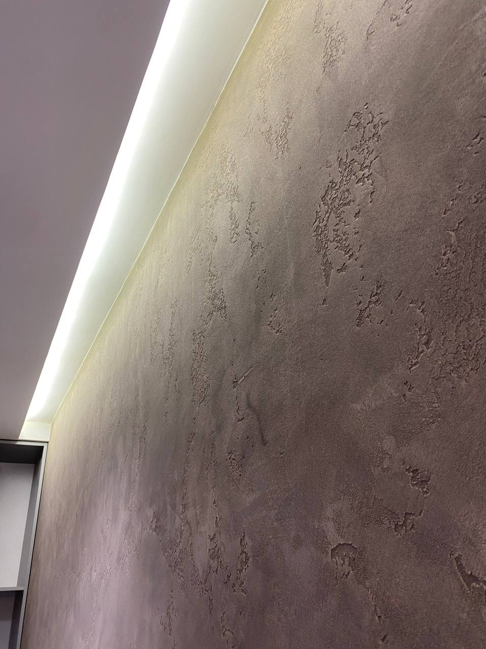 Teksturowana ściana w odcieniach brązu z subtelnym oświetleniem LED wzdłuż sufitu, ukazująca detale wykończenia i nieregularną fakturę powierzchni.