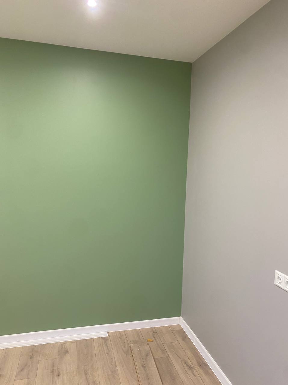 Pomieszczenie po odświeżeniu: ściana w odcieniu zieleni, kontrastująca z szarą ścianą narożną, jasna podłoga z paneli, biała listwa przypodłogowa i gniazdko elektryczne w prawym dolnym rogu.