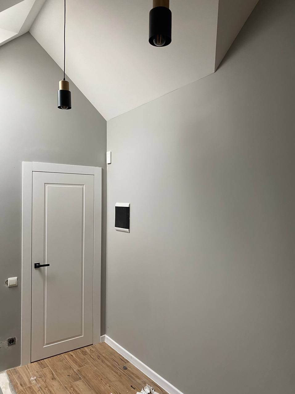 Świeżo pomalowane wnętrze w odcieniach szarości z drewnianą podłogą i nowoczesnym oświetleniem sufitowym. Widoczna biała stolarka drzwiowa z czarną klamką oraz panel sterowania inteligentnego domu...