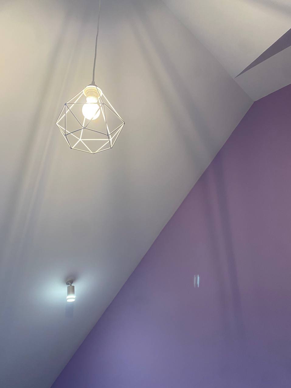 Nowoczesna lampa wisząca z geometrycznym kloszem, oświetlająca biały sufit i liliową ścianę w skosie, widoczna także mała lampa sufitowa.