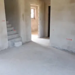 Surowe wnętrze domu po tynkowaniu maszynowym, widoczne schody w stanie surowym, ściany z naniesionym tynkiem, okno w tle i widoczne ślady po instalacjach elektrycznych.