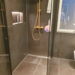 Nowoczesna kabina prysznicowa z ciemnymi płytkami, złotą armaturą i wnęką na kosmetyki. Widoczny odpływ liniowy i szklana ściana kabiny.