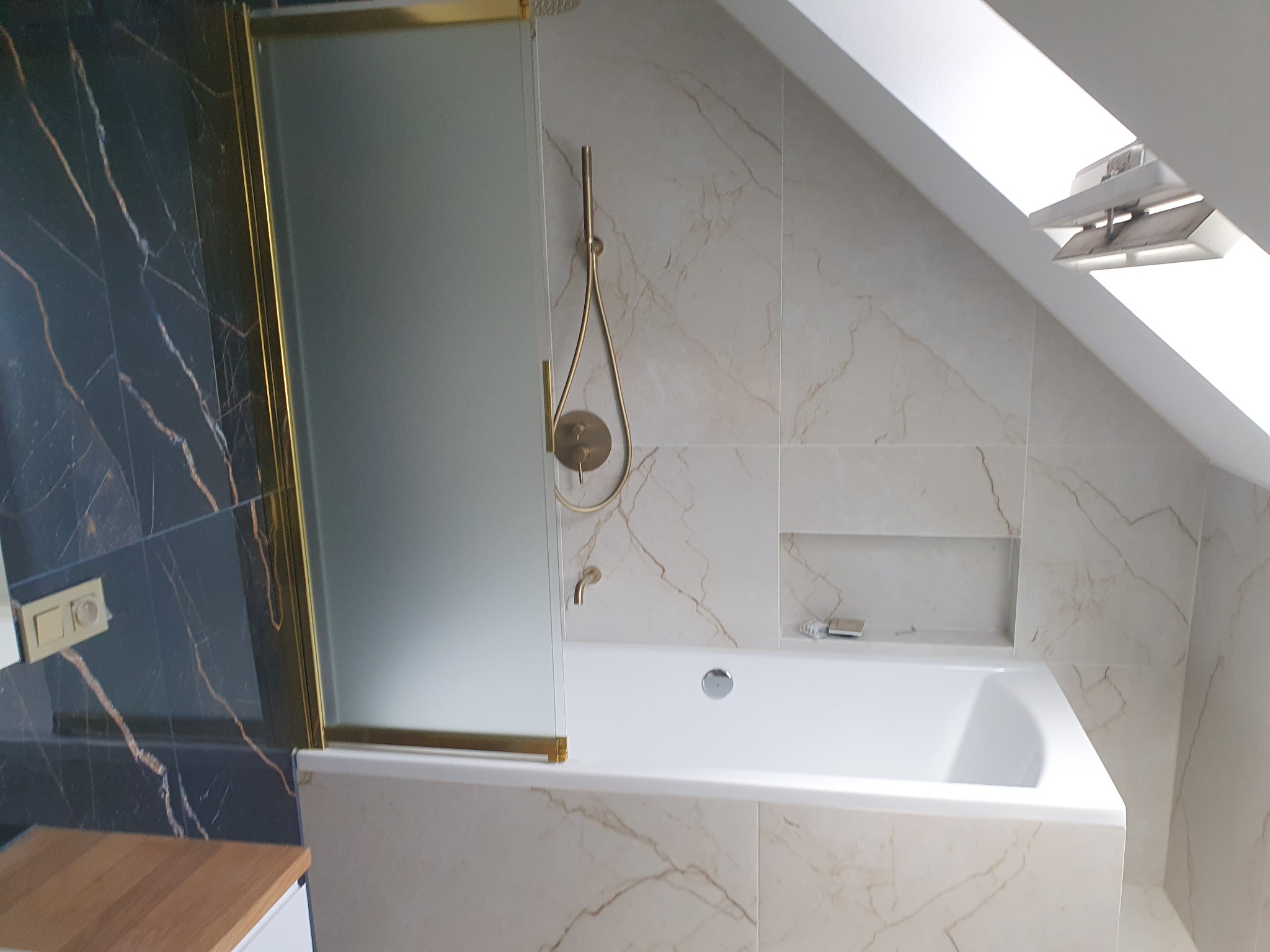 Elegancka łazienka na poddaszu z wanną, złotą armaturą i czarnymi płytkami imitującymi marmur. Widoczne okno dachowe i wnęka w ścianie.