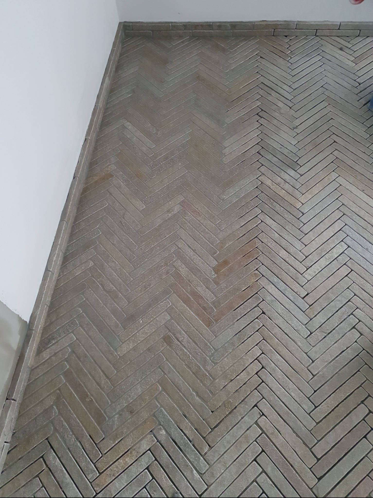 Ukończona podłoga z płytek ułożonych w jodełkę, przy białej ścianie. Widoczne detale fug i krawędzi płytek. Jasne, naturalne światło.