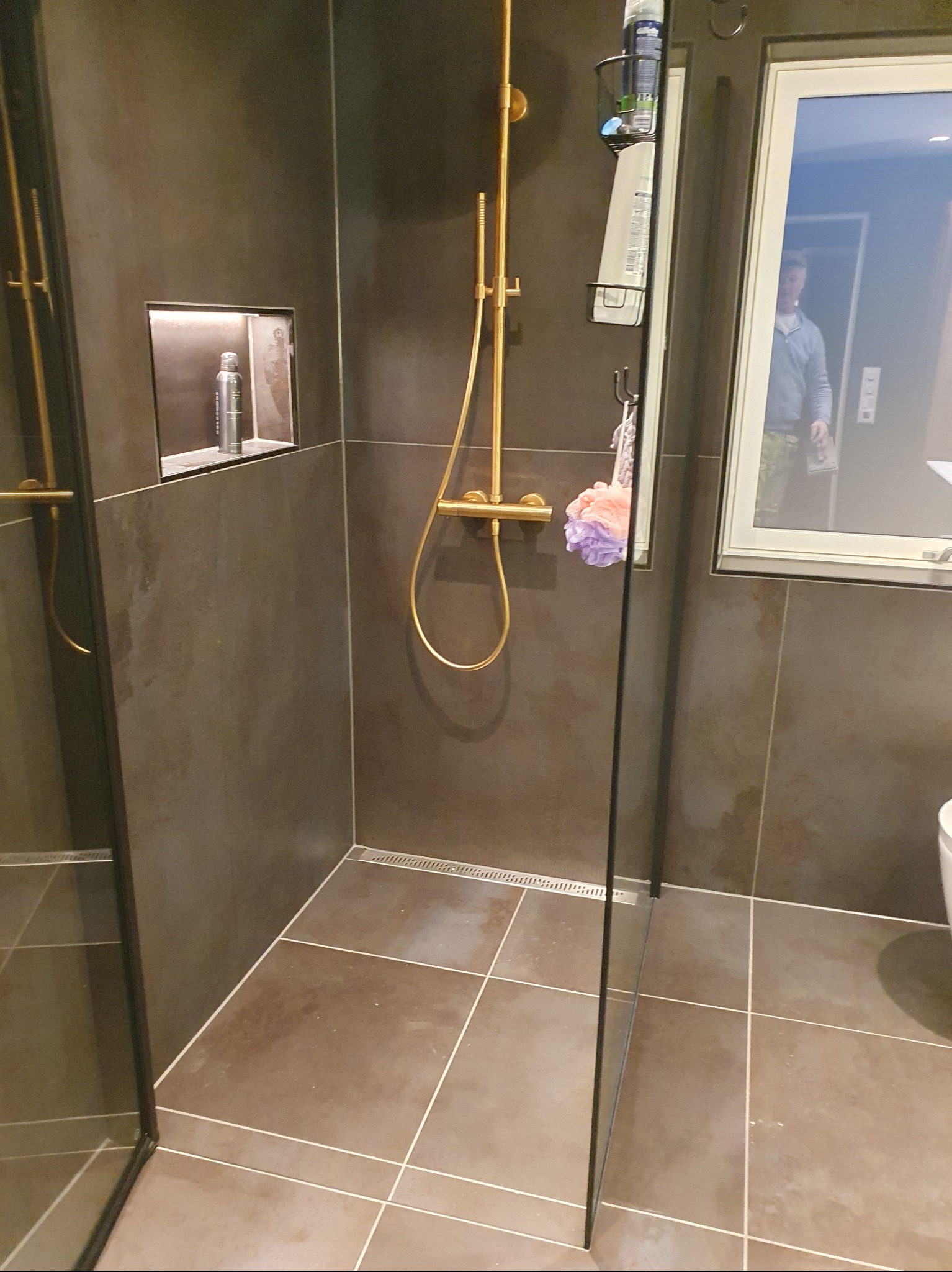 Nowoczesna kabina prysznicowa z ciemnymi płytkami, złotą armaturą i wnęką na kosmetyki. Widoczny odpływ liniowy i szklana ściana kabiny.