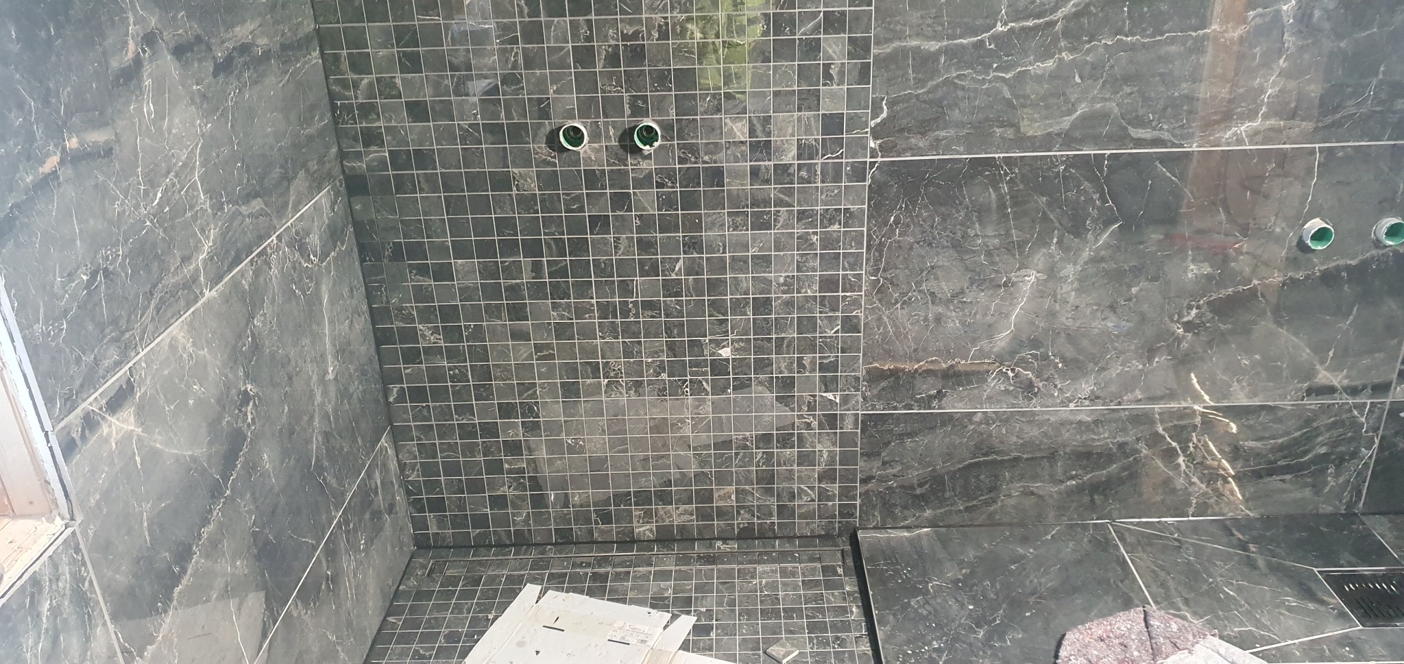 Częściowo wykończona łazienka z płytkami imitującymi marmur i mozaiką. Widoczne wyprowadzenia wodne i odpływ liniowy. Prace glazurnicze w toku.