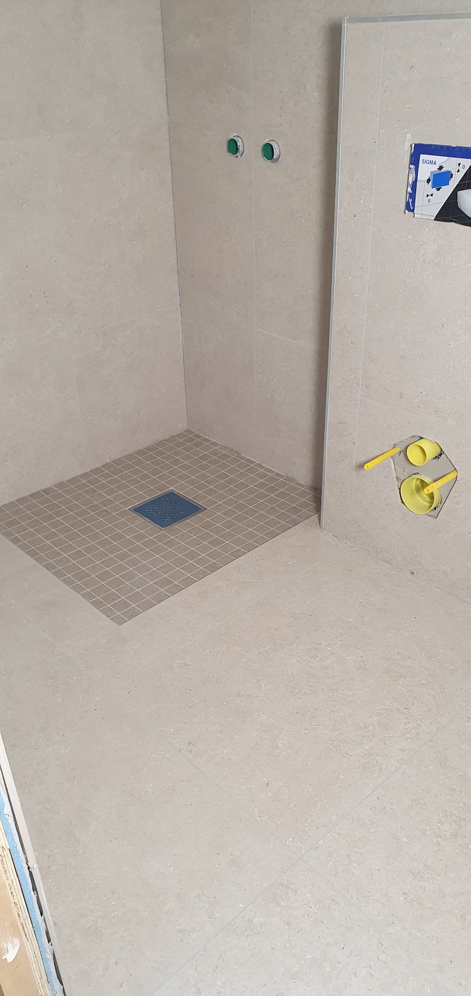 Beżowa łazienka w trakcie wykańczania: kwadratowy odpływ liniowy z mozaiki na podłodze, widoczne instalacje wodne i fragment narzędzi do montażu płytek.