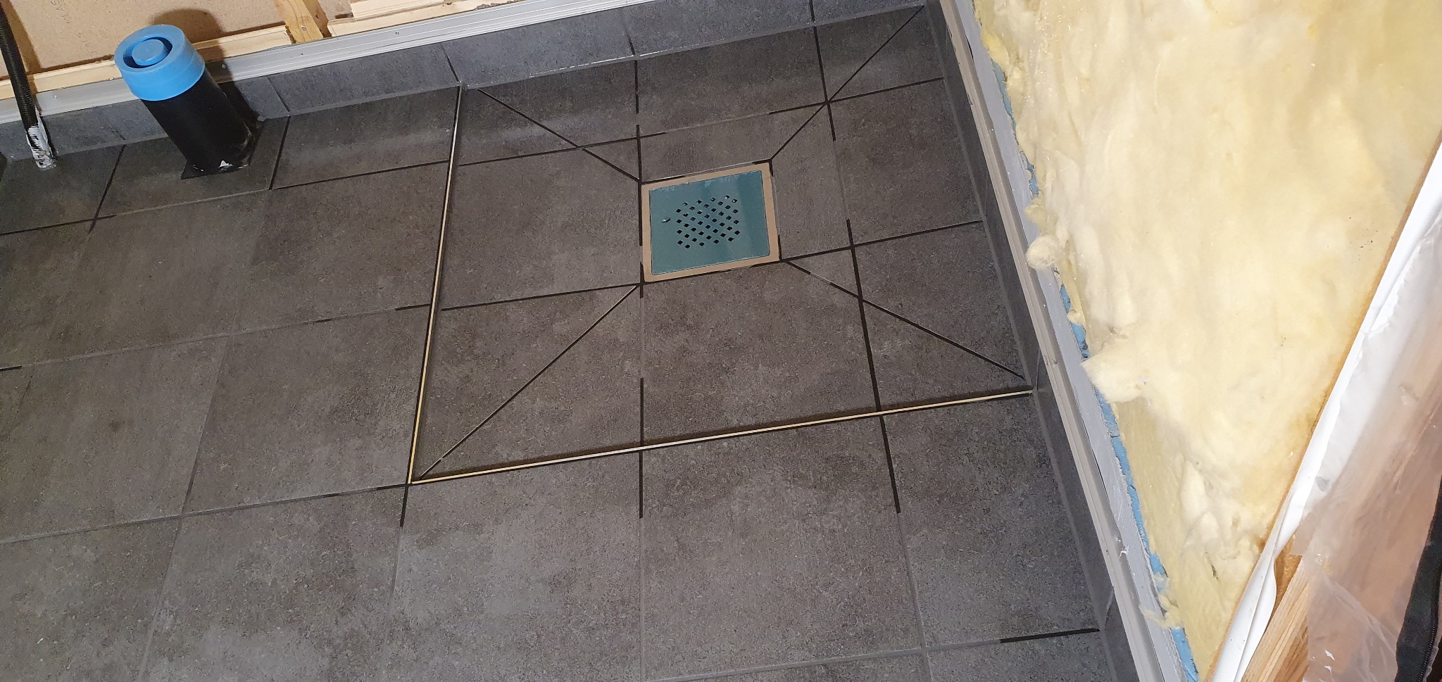 Ułożone ciemne płytki podłogowe z odpływem liniowym w kabinie prysznicowej, widoczne profile i izolacja ściany. Detal wykończenia łazienki.