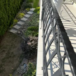 Widok z balkonu na kamienne schody w ogrodzie, otoczone zielenią, z dekoracyjną metalową balustradą rzucającą cienie na jasną podłogę balkonu.