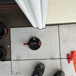 Adrian Turecki - Układanie jasnych płytek podłogowych przy użyciu przyssawki Raimondi i krzyżyków dystansowych, widoczne zabrudzone spodnie i buty montera.