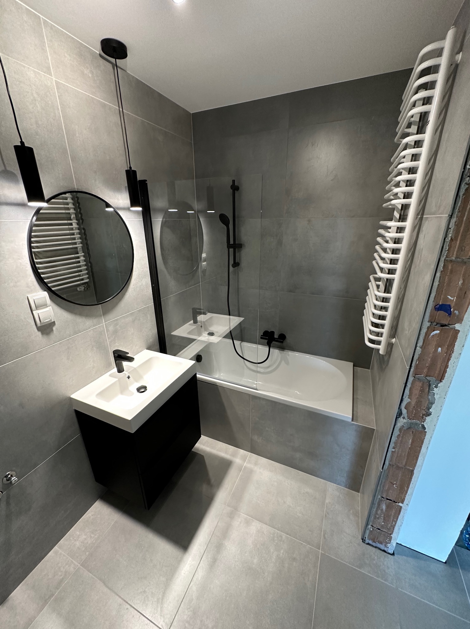 Nowoczesna łazienka w odcieniach szarości z czarnymi akcentami, okrągłe lustro z podświetleniem LED, biała umywalka nablatowa na czarnej szafce, grzejnik drabinkowy i widoczny fragment surowej...
