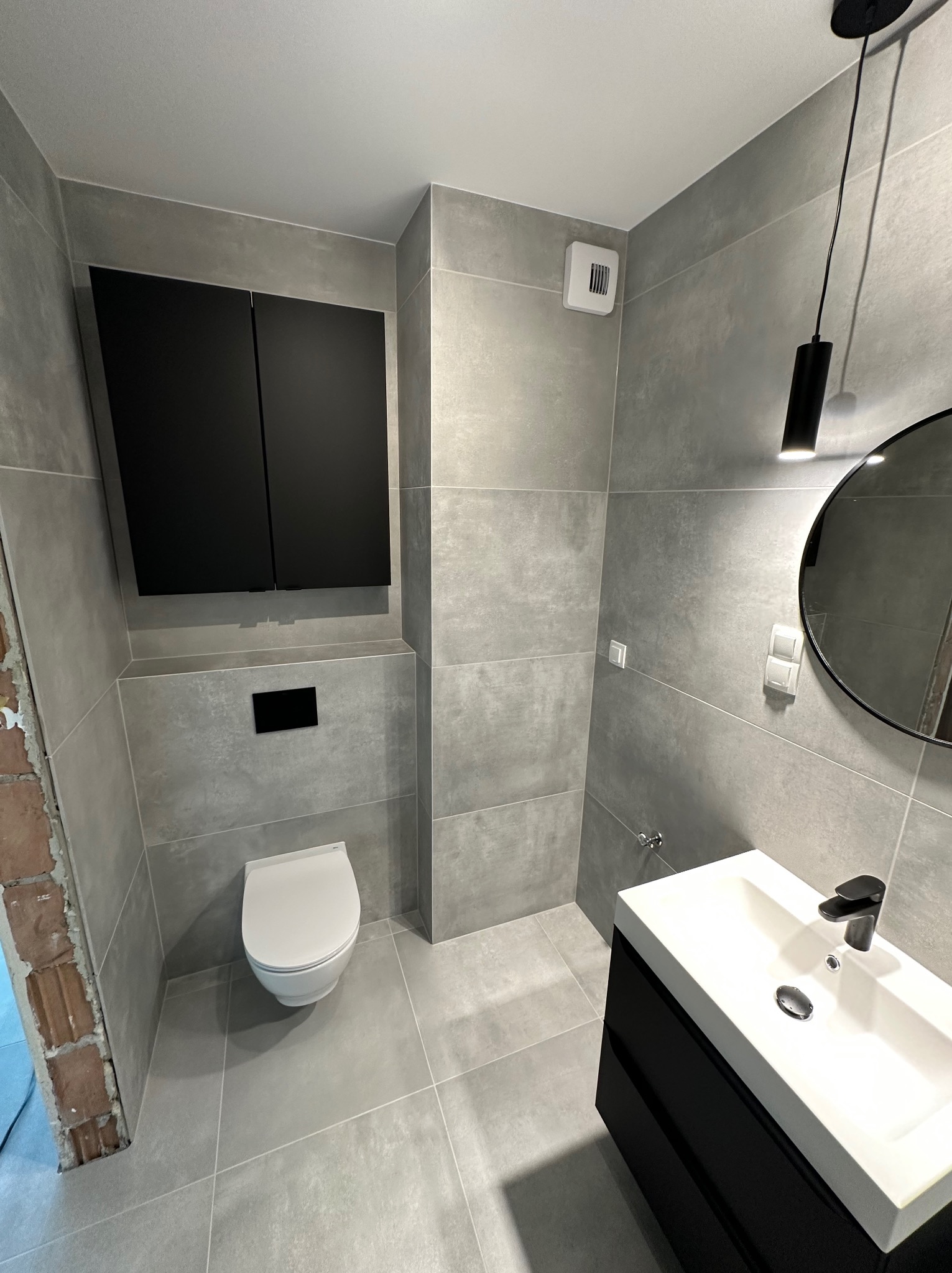 Nowoczesna łazienka z szarymi płytkami, wiszącą toaletą i czarną szafką pod umywalką, oświetlona czarną lampą wiszącą; widoczny fragment surowej ściany przed wykończeniem.