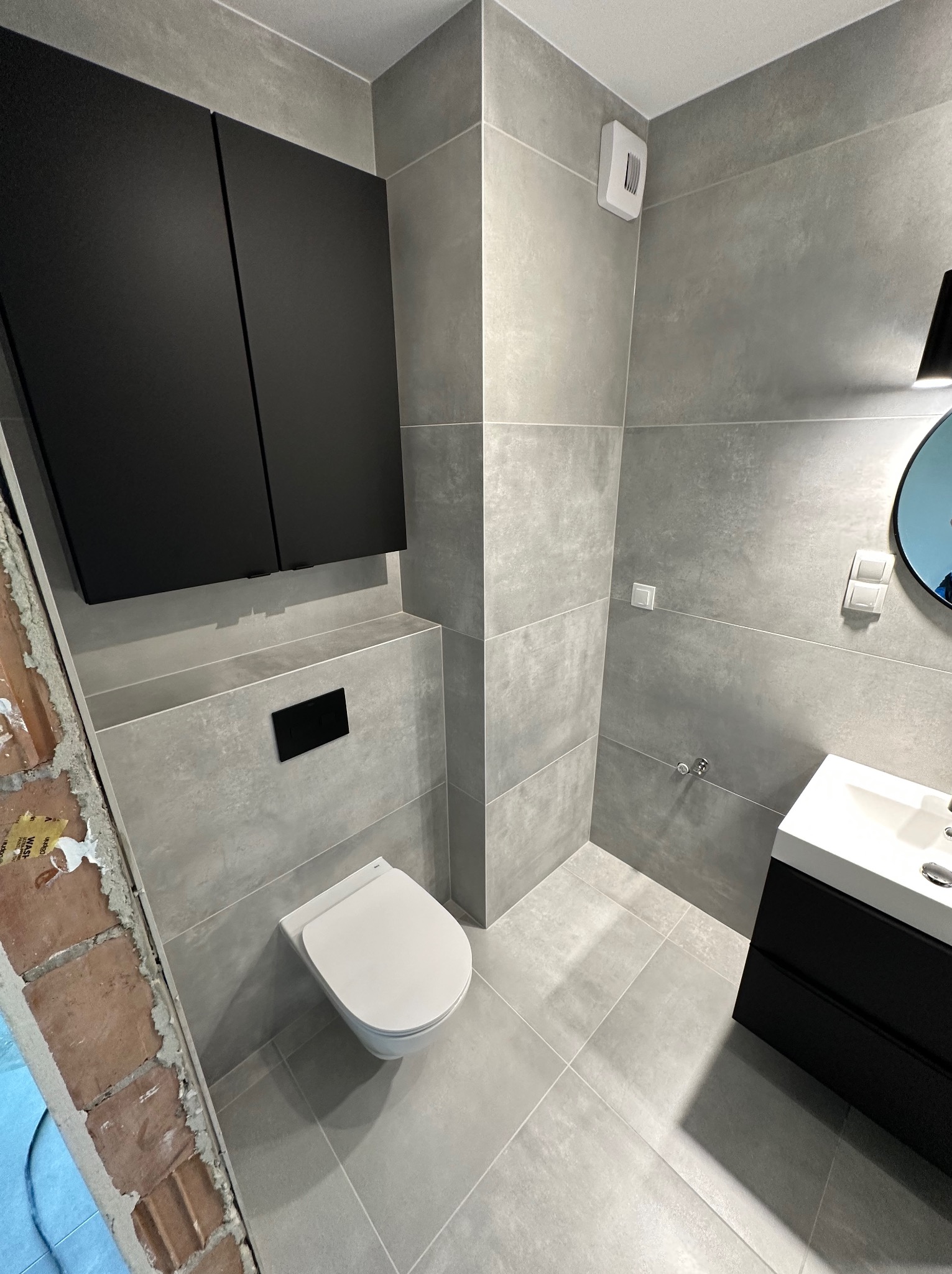 Nowoczesna łazienka z szarymi płytkami, czarnymi szafkami wiszącymi i umywalką; widoczny fragment ściany z cegły sugerujący etap prac.