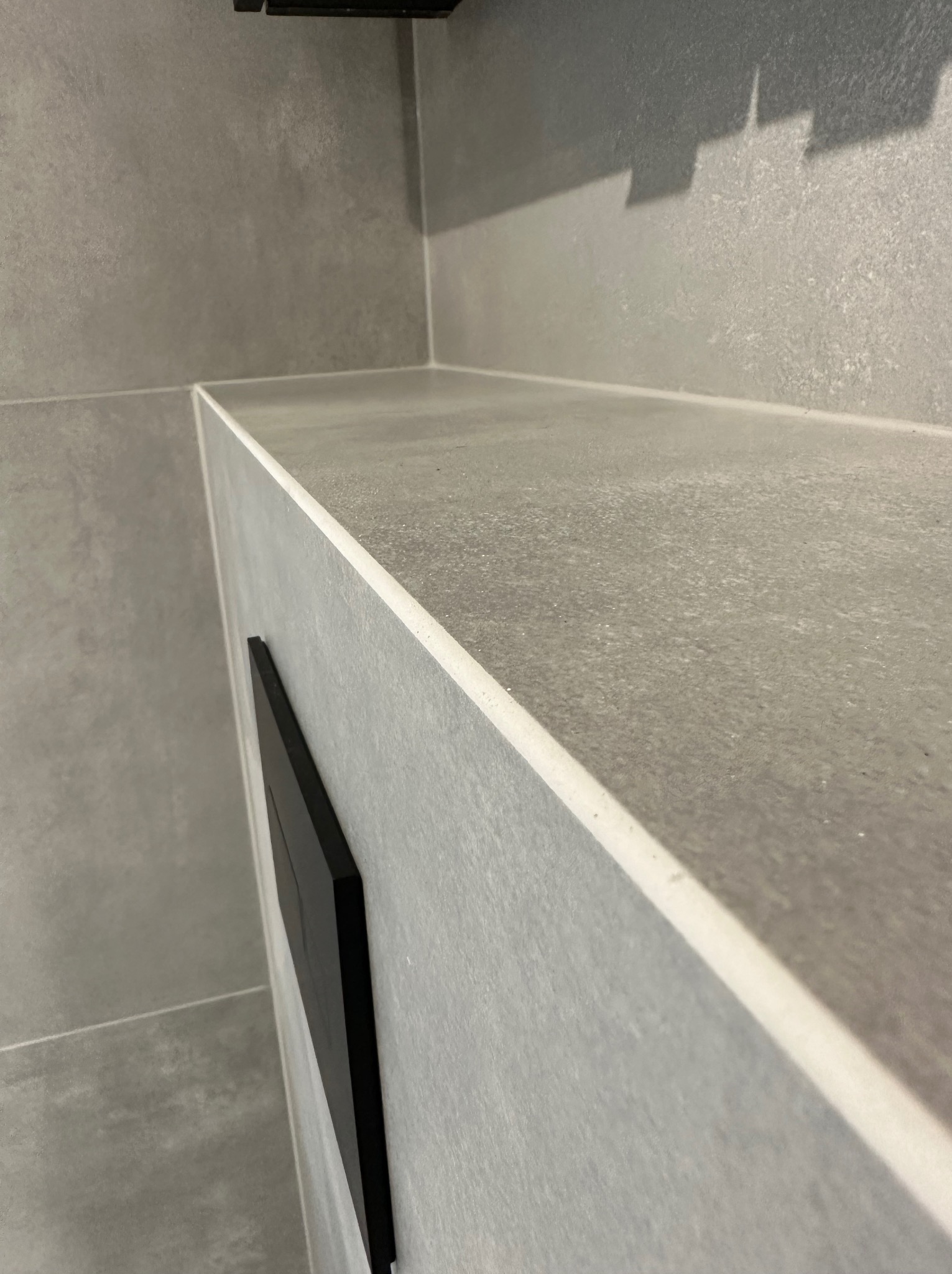 Szara, minimalistyczna łazienka z blatem z płytek imitujących beton, czarny panel sterowania spłuczką, widok z góry i boku.