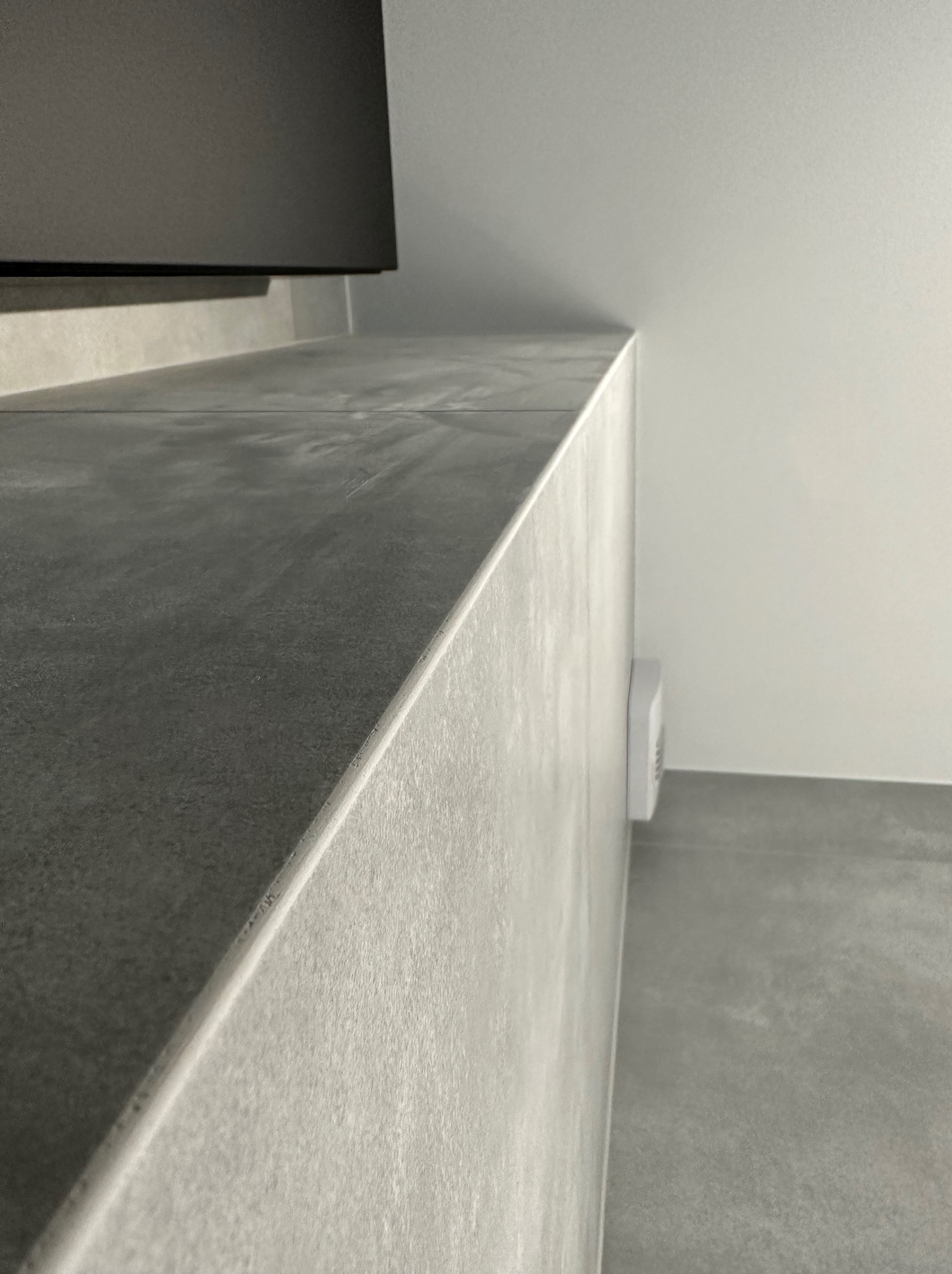 Szara, minimalistyczna łazienka z płytkami imitującymi beton, widoczny fragment blatu umywalkowego i wentylator ścienny.