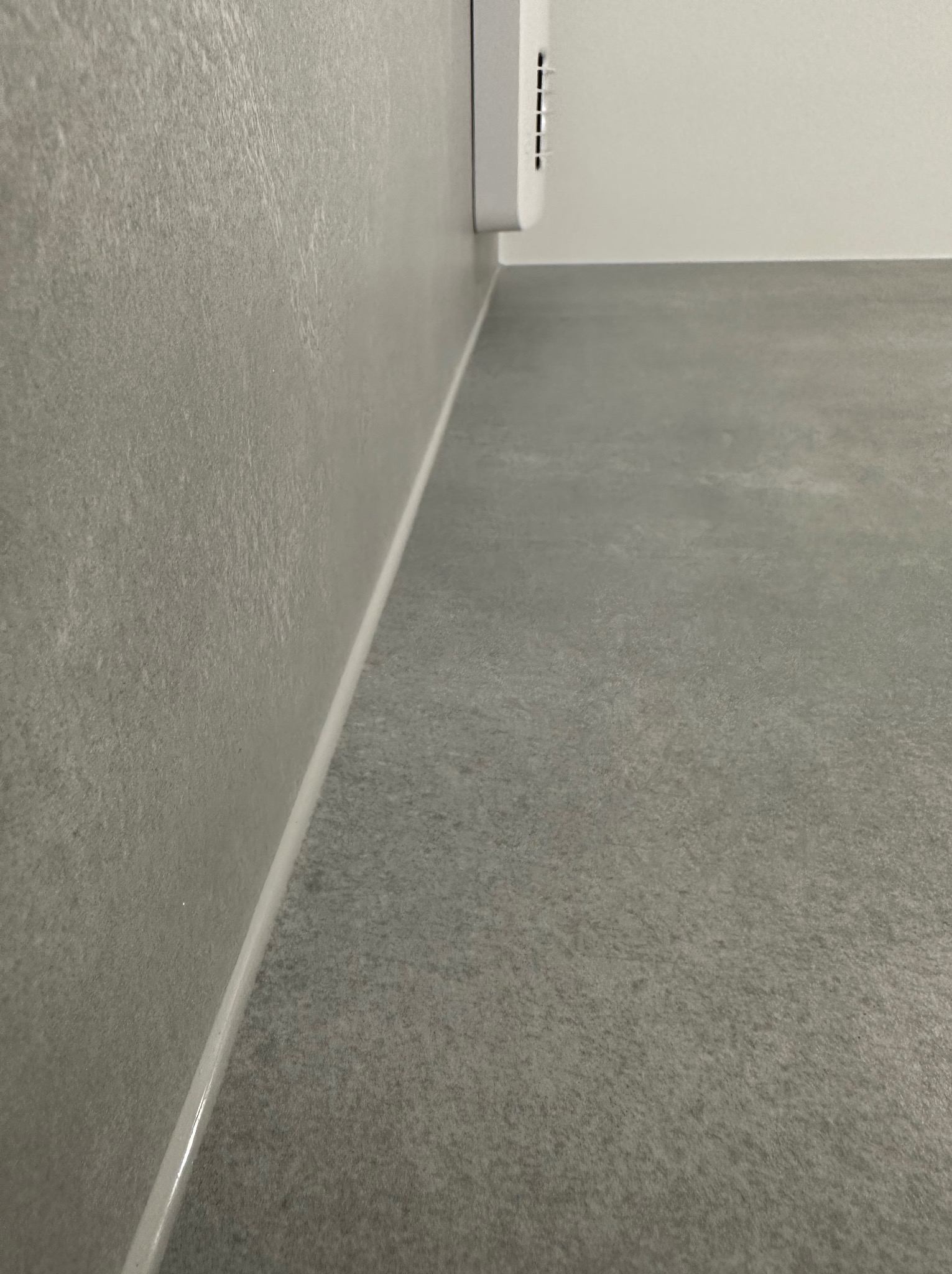 Szara ściana z fakturą i szara podłoga, wykończone białą listwą przypodłogową, z widocznym fragmentem białego urządzenia na ścianie.