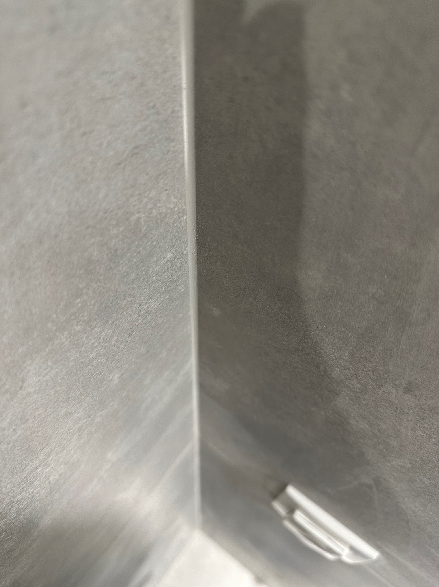 Szare płytki ceramiczne na ścianie w rogu pomieszczenia, widoczna listwa wykończeniowa i fragment uchwytu na ręcznik.