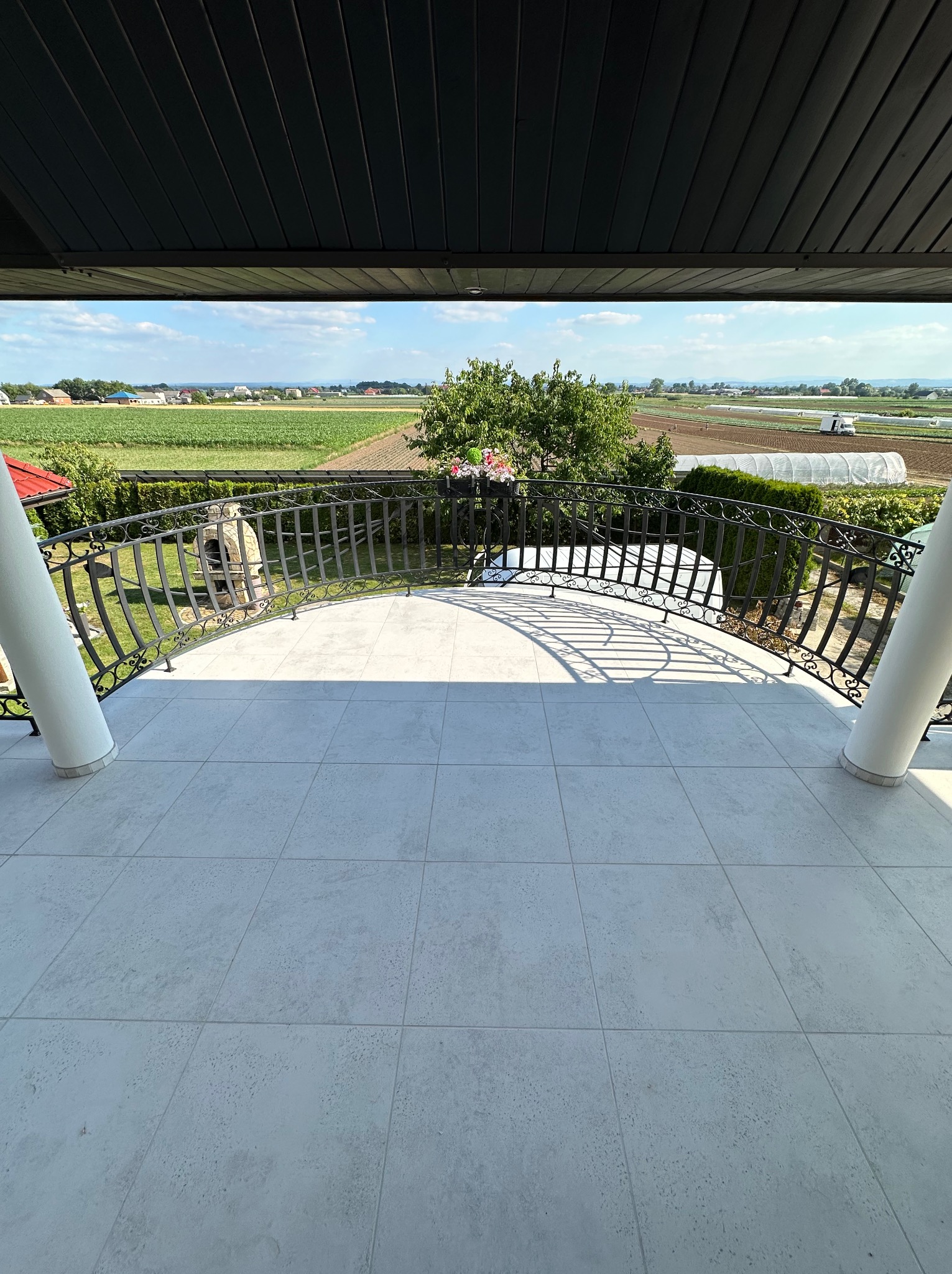 Biała, ceramiczna podłoga balkonu z kutą, czarną balustradą i widokiem na wiejski krajobraz z uprawami i szklarnią w tle.