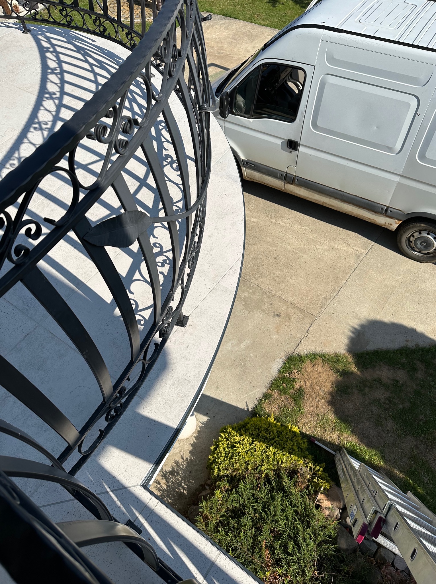 Kuta, czarna balustrada balkonu z dekoracyjnymi elementami, jasne płytki na posadzce, biały samochód dostawczy w tle oraz rozłożona drabina oparta o krzewy.