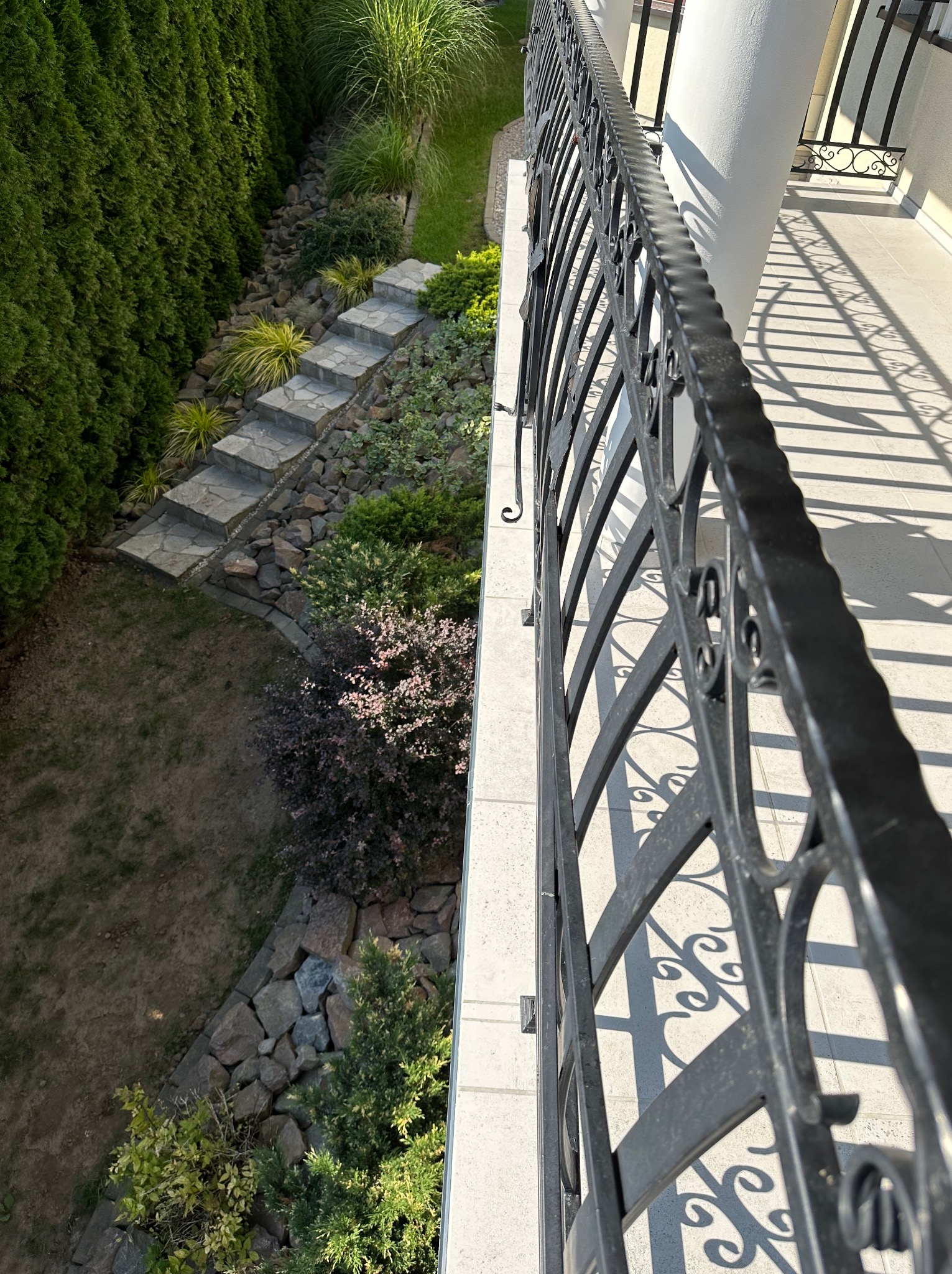 Widok z balkonu na kamienne schody w ogrodzie, otoczone zielenią, z dekoracyjną metalową balustradą rzucającą cienie na jasną podłogę balkonu.