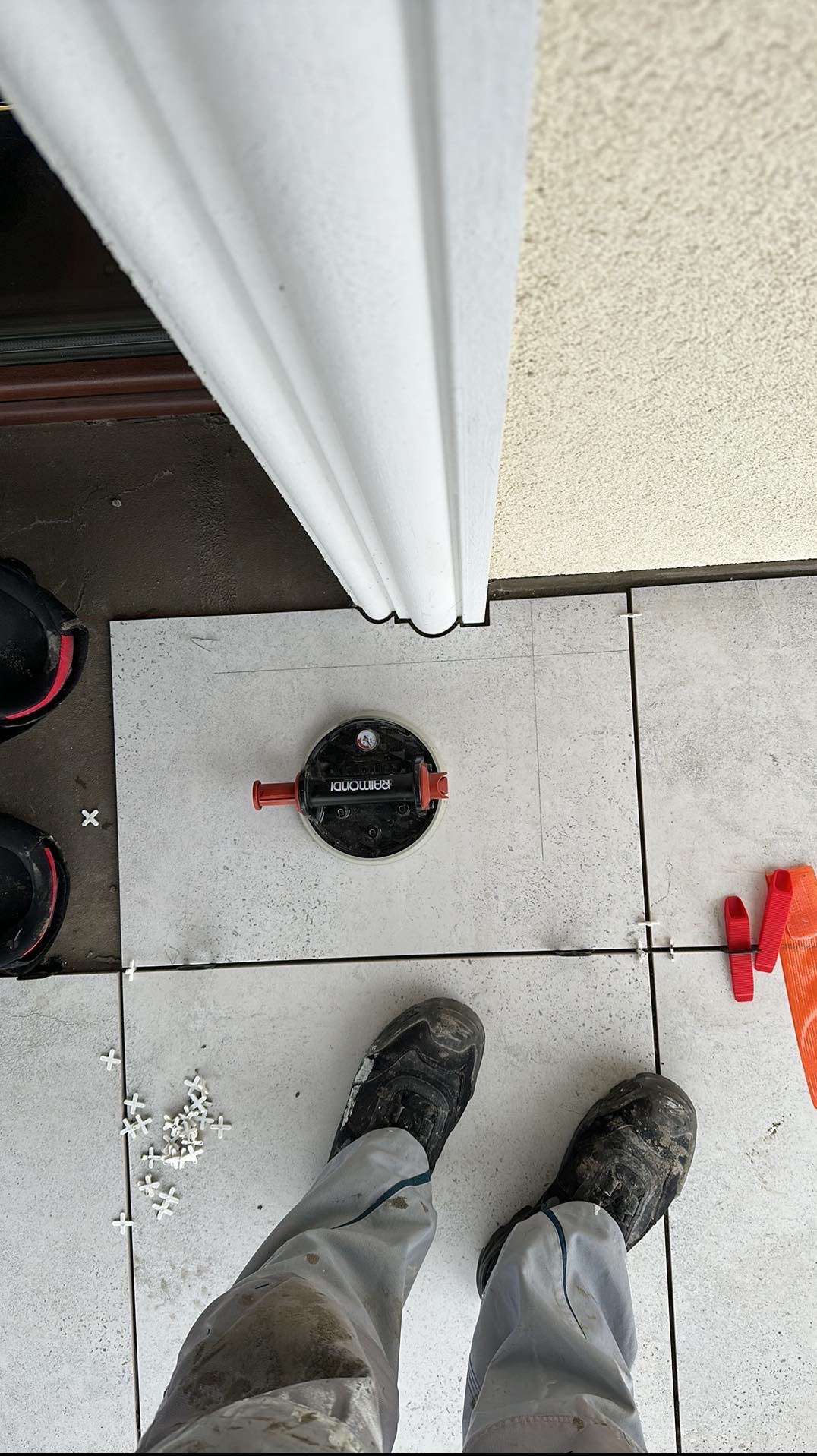 Układanie jasnych płytek podłogowych przy użyciu przyssawki Raimondi i krzyżyków dystansowych, widoczne zabrudzone spodnie i buty montera.