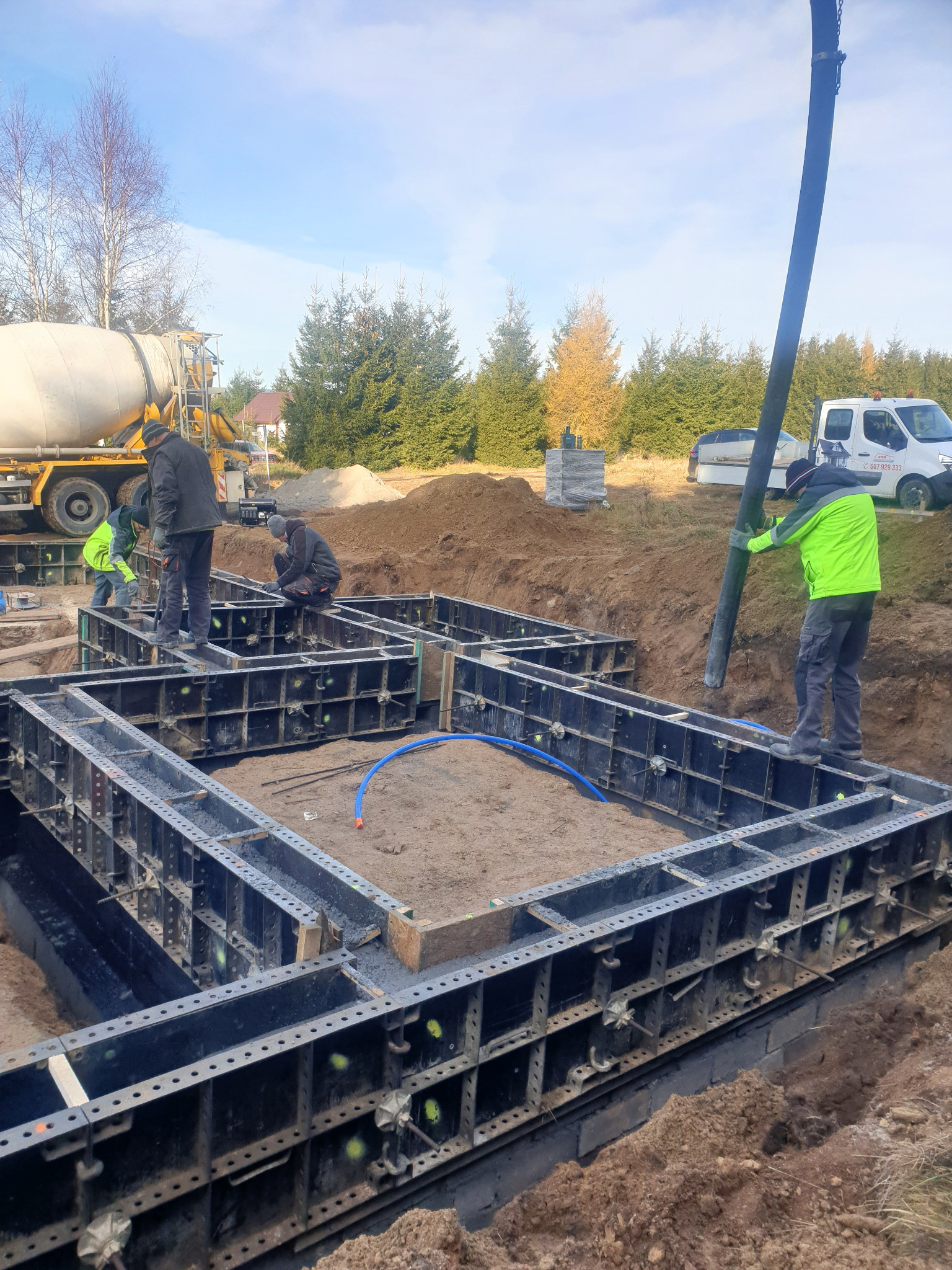 Wylewanie betonu do szalunków fundamentowych przez pracowników w odblaskowych kamizelkach, z betonomieszarki i pompy do betonu na tle jesiennego krajobrazu.
