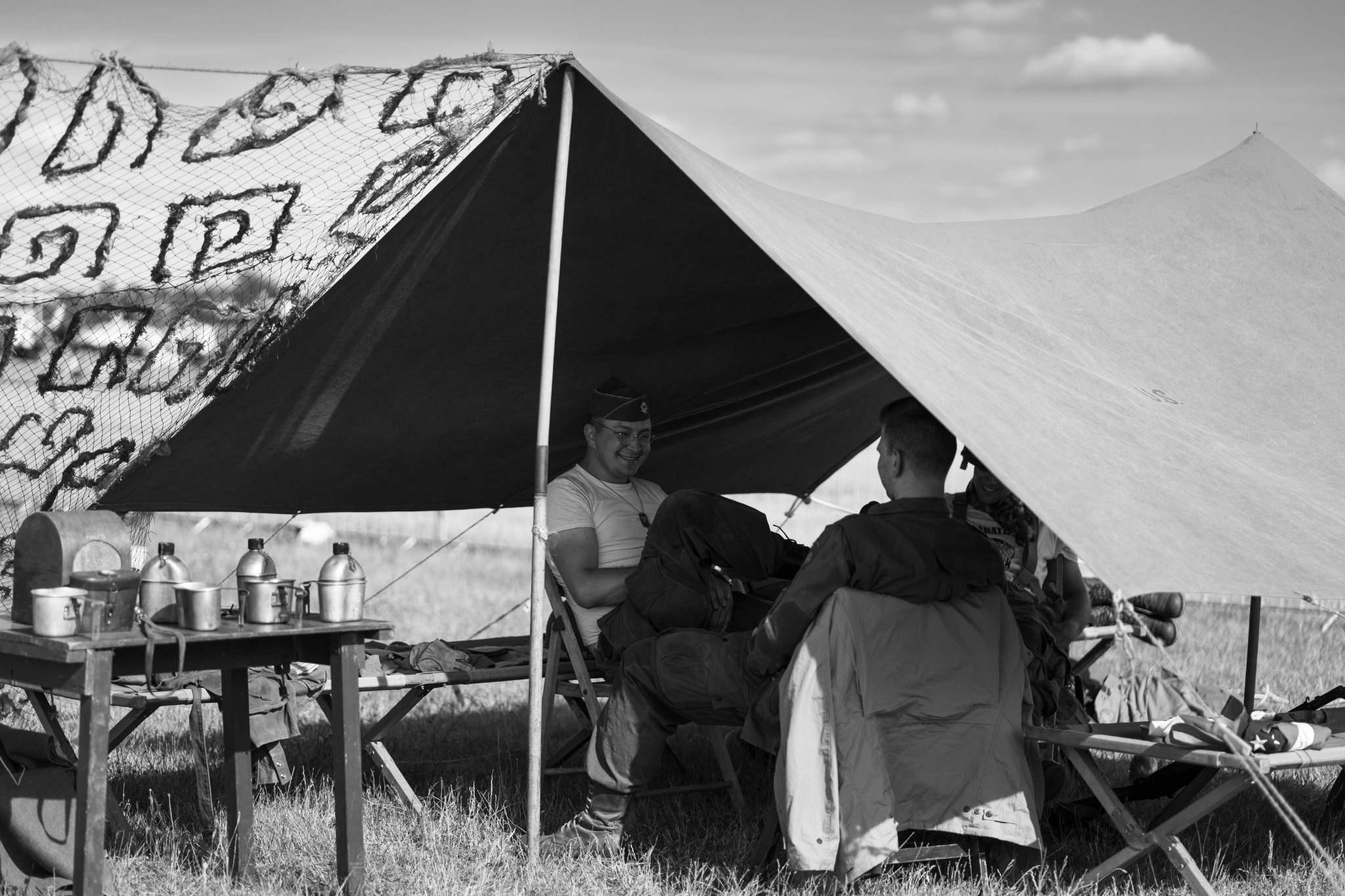 Czarno-białe zdjęcie przedstawia dwóch mężczyzn w strojach historycznych siedzących pod namiotem, na stole obok metalowe naczynia, a na zewnątrz trawa i niebo.