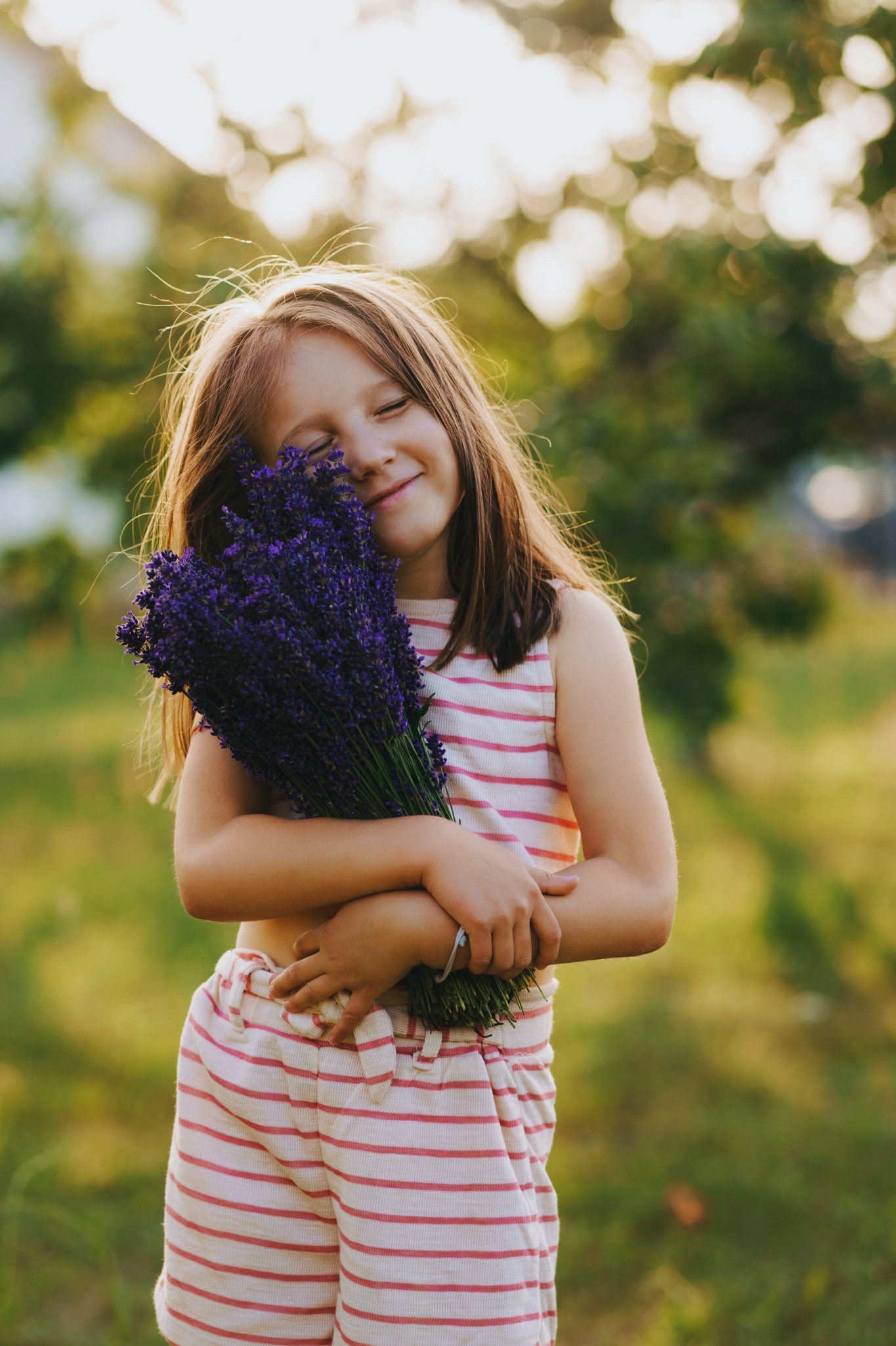 Uśmiechnięta dziewczynka w paski obejmuje bukiet świeżej lawendy na tle rozmytej zieleni.