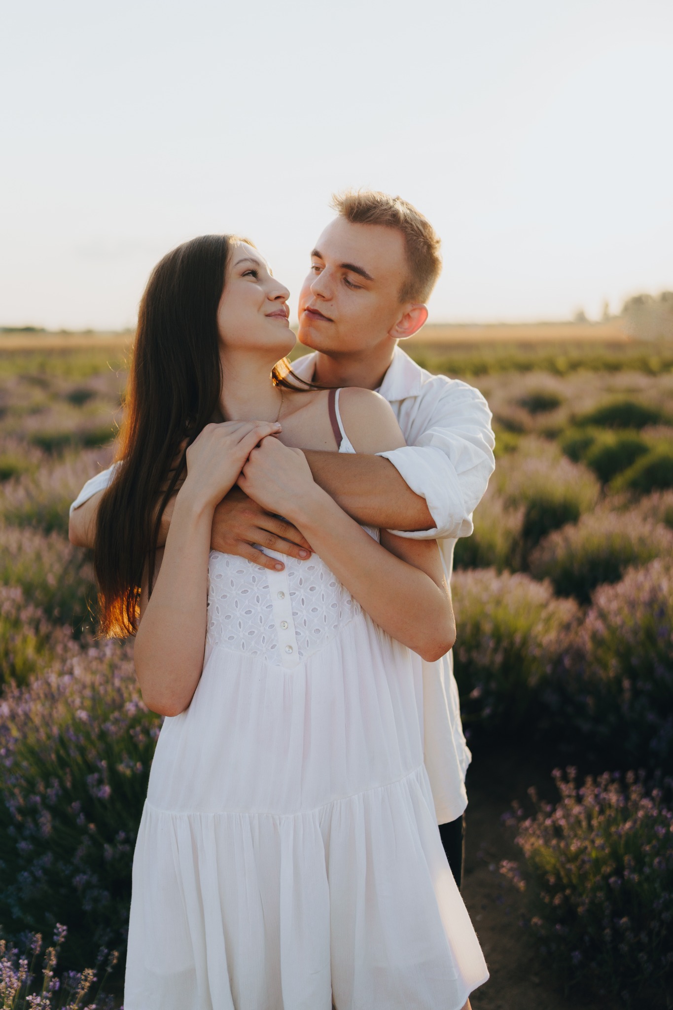 Uśmiechnięta para w białych ubraniach obejmuje się na tle kwitnącego pola lawendy podczas sesji zdjęciowej w plenerze, w ciepłym, naturalnym świetle zachodzącego słońca.
