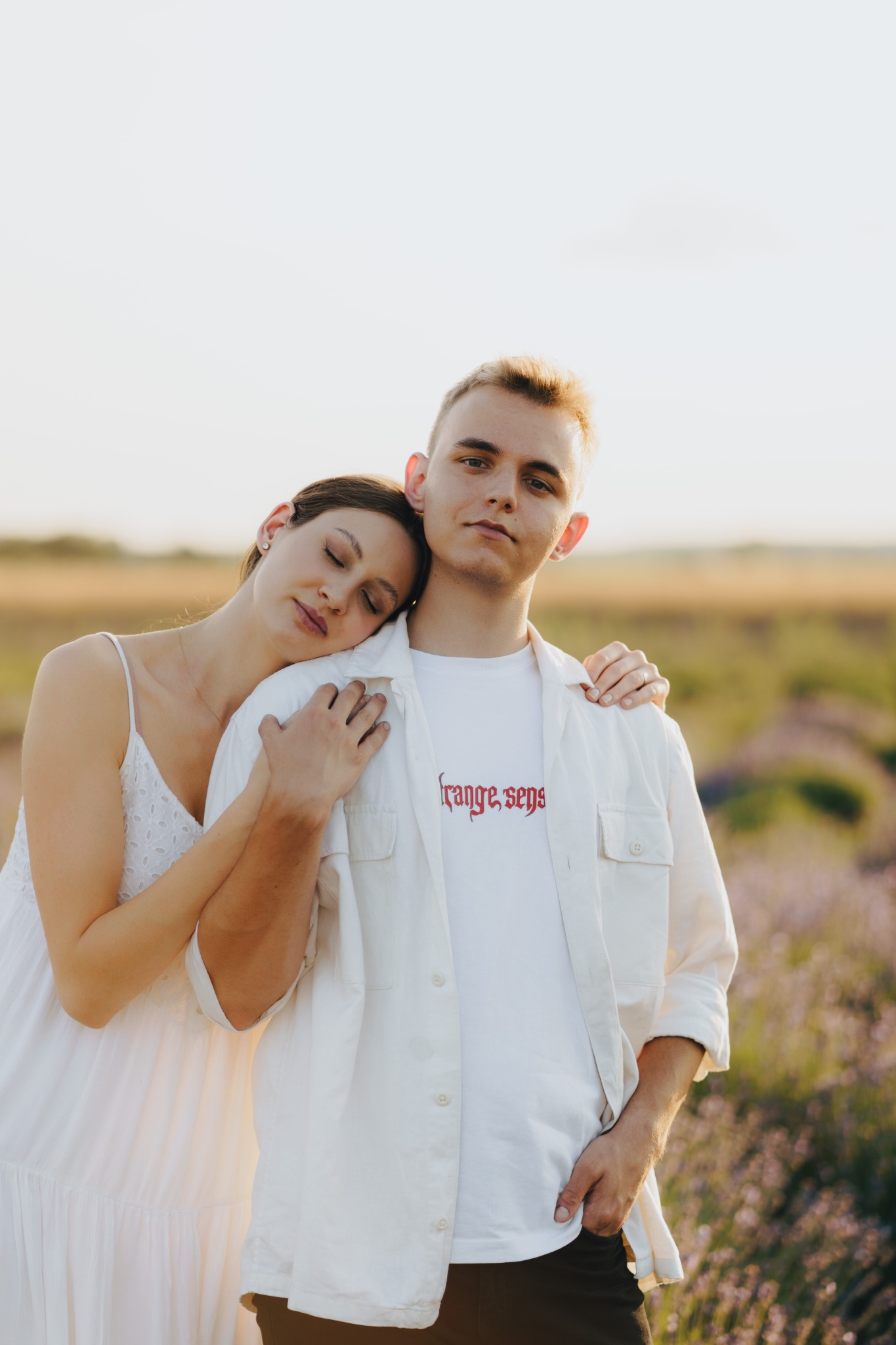 Romantyczna sesja plenerowa: kobieta w białej sukience opiera głowę na ramieniu mężczyzny w białej koszuli z czerwonym napisem, w tle pole lawendy w miękkim, zachodzącym słońcu.