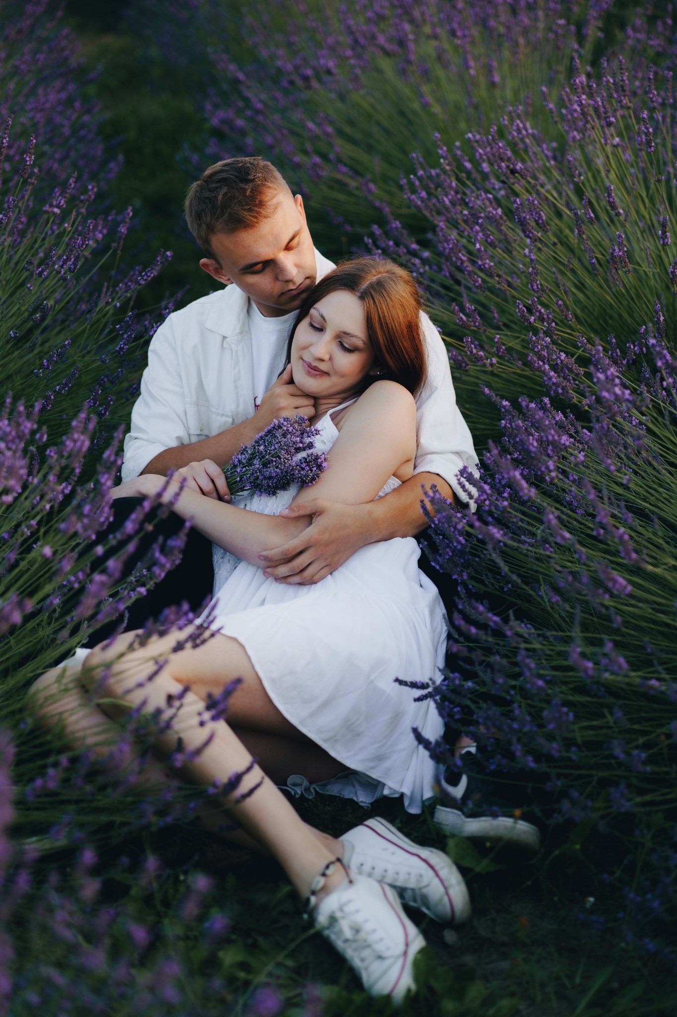 Para w białych ubraniach siedzi w polu lawendy, mężczyzna obejmuje kobietę trzymającą bukiet lawendy.