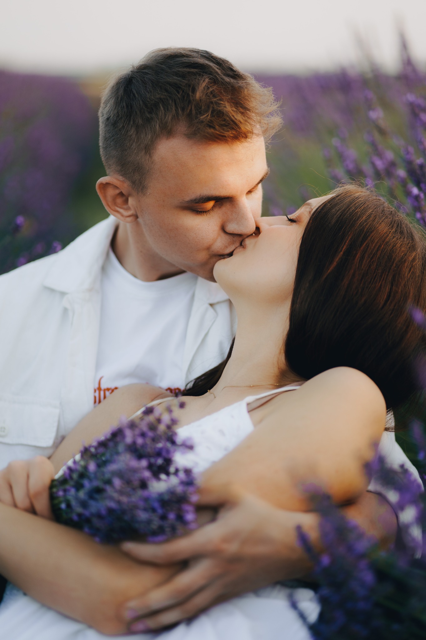 Zakochana para całuje się na polu lawendy, kobieta trzyma bukiet lawendy, ciepłe światło.